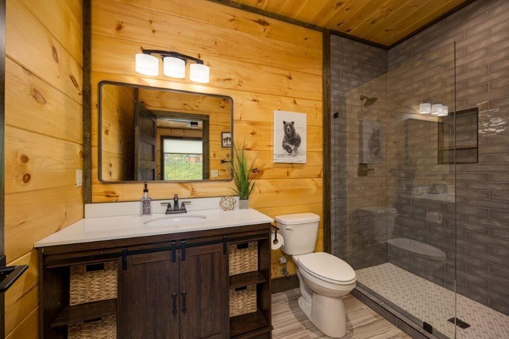 Une salle de bain rustique, avec une cabine en bois, un lavabo blanc, des meubles en bois foncé, des toilettes blanches et une douche à parois de verre.