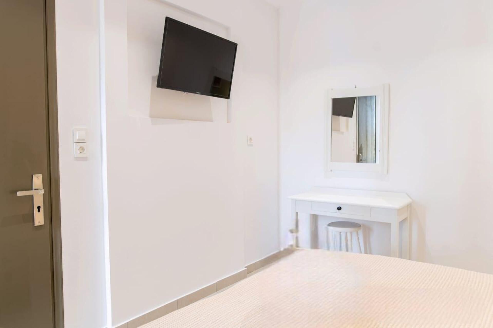 Et hotelværelse med en seng, et fjernsyn ophængt på væggen, et lille hvidt toiletbord med spejl og en dør med nøglelås.