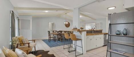 Una sala de estar moderna con un sofá, una mesa de centro y una alfombra. Hay un comedor con una mesa y sillas. La cocina cuenta con un refrigerador, estantes y una encimera.
