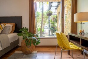 A room with a bed, a large potted plant, a yellow chair, a table with a lamp, and a view of greenery outside.
