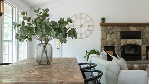Een woonkamer met een stenen openhaard, een klok aan de muur en een vaas met groene planten op een houten tafel.