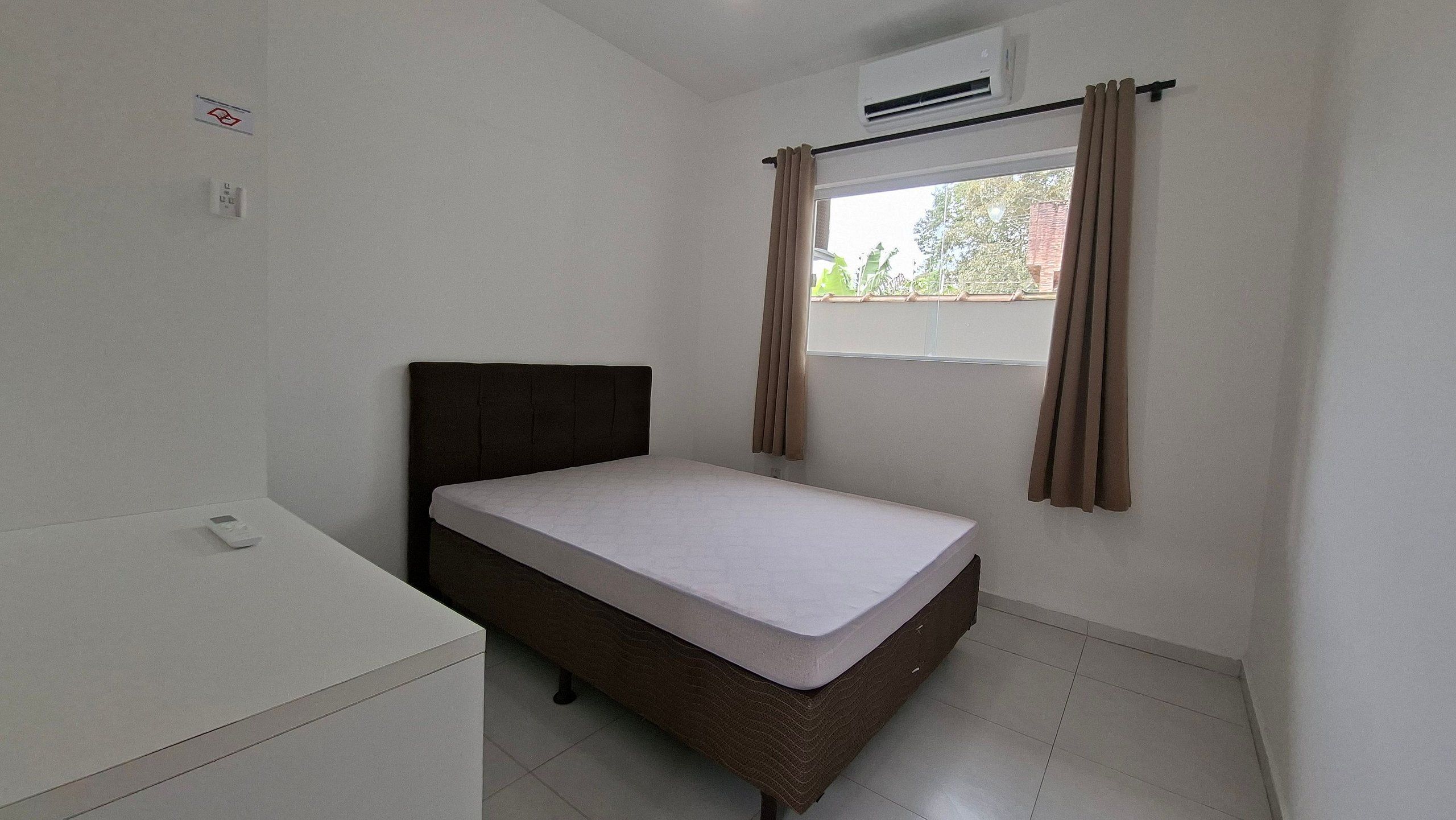 Quarto pequeno com cama de solteiro, colchão branco, cabeceira marrom e janela com cortinas.