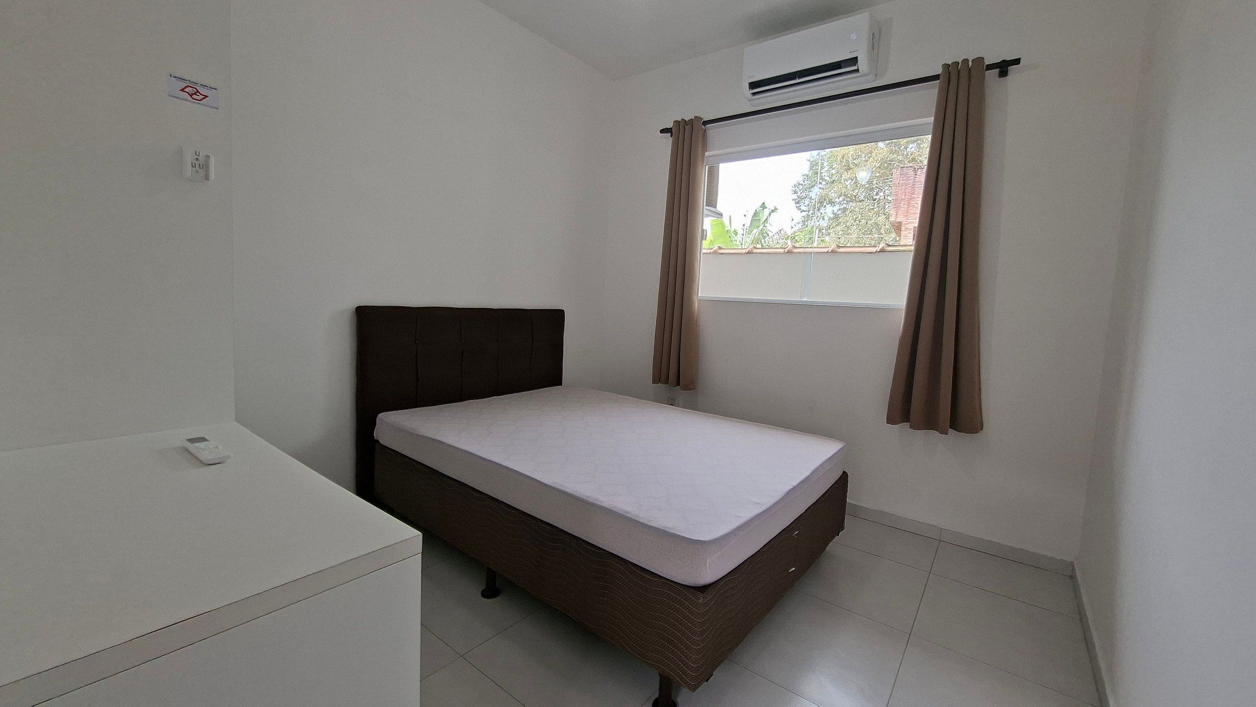 A small, single-bed room with a plain white mattress, a dark headboard, and a window with brown curtains.