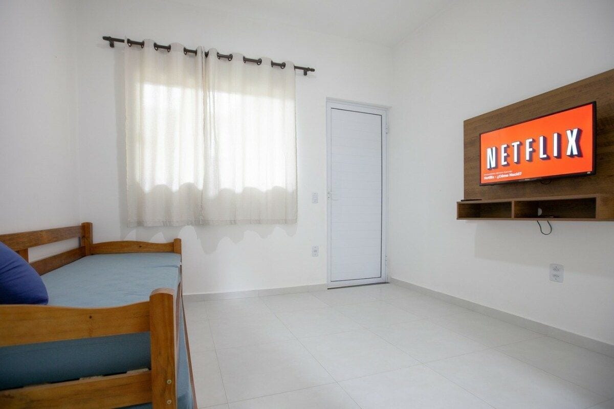 Una habitación con cama, una puerta, una ventana con cortinas y un televisor de pared mostrando el logotipo de Netflix.