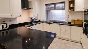 A modern kitchen with white cabinets, a black countertop, and a window with blinds. - South Bay Beach House (Bridlington)