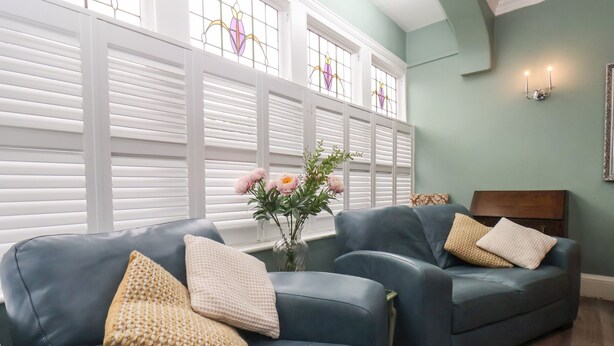 A room with two dark blue leather sofas, each with a textured throw pillow, positioned in front of a window with white shutters. A vase with pink flowers is placed on the windowsill. - South Bay Beach House (Bridlington)