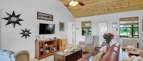 A living room with a wooden ceiling, a ceiling fan, a TV, a bookshelf, a sofa, and a dining table with chairs.