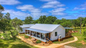 A modern house with a covered porch and a deck, surrounded by lush greenery and a clear sky. - Luxurious Cottage on 3-acre's in Fred Wine Country (Fredericksburg)