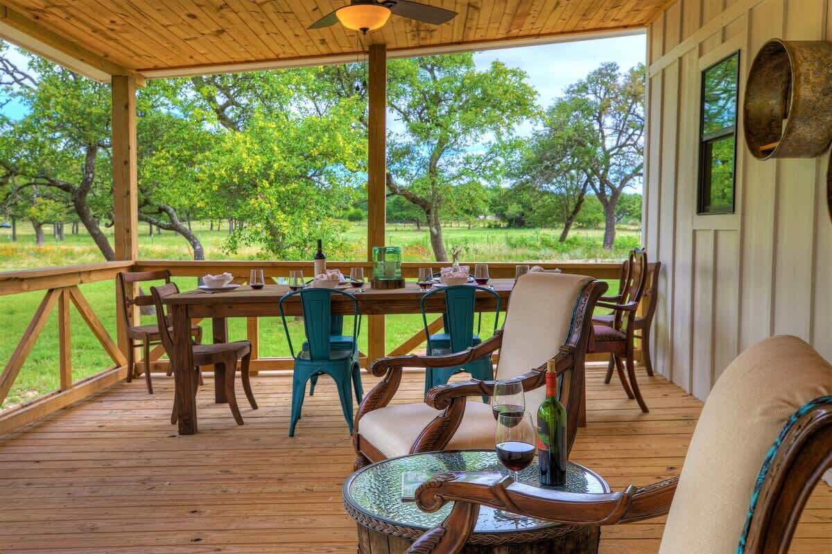 A covered outdoor dining area with a wooden table and chairs, a glass table, and a view of a green landscape with trees.