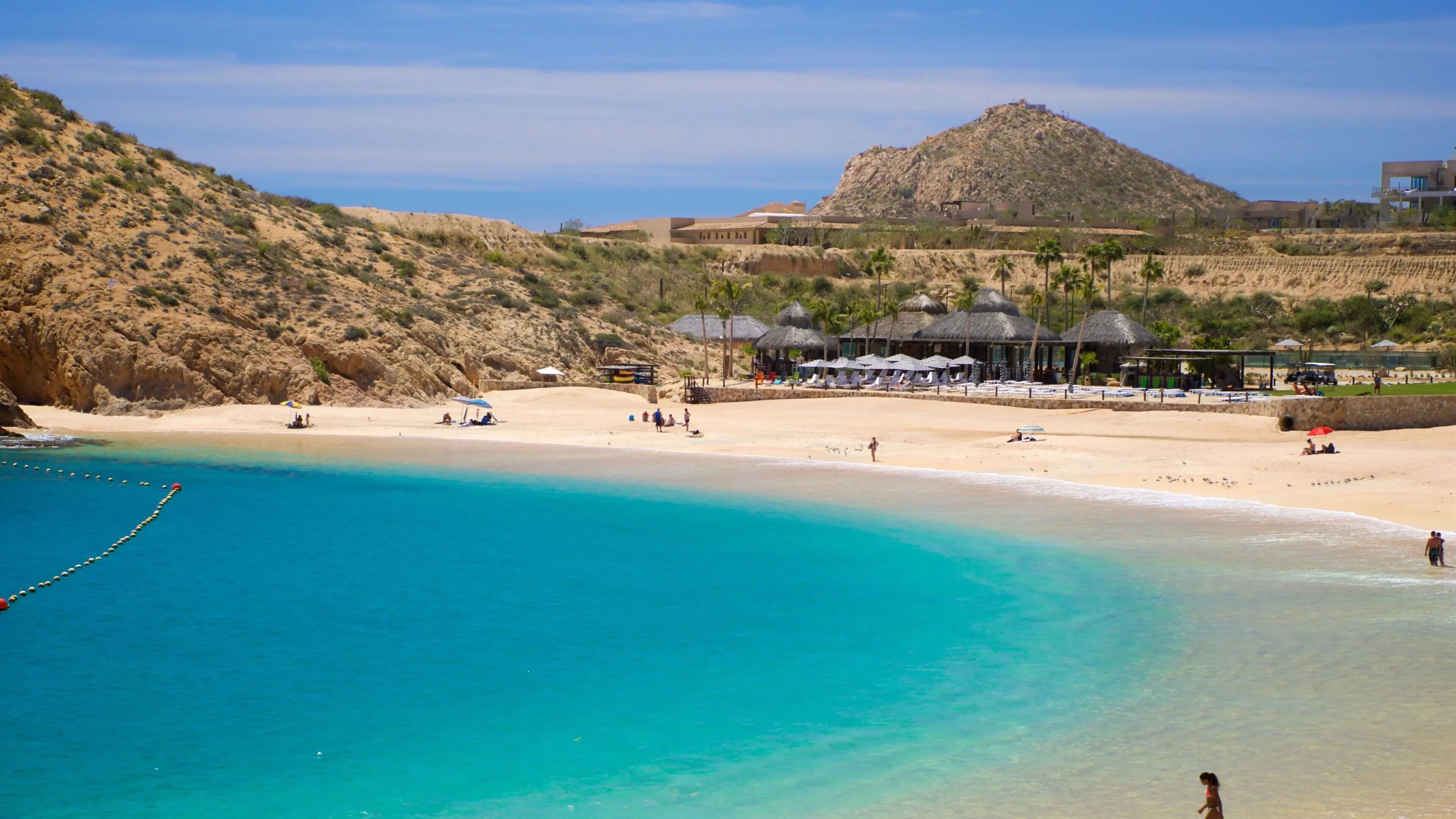 A beach resort with clear turquoise waters, a sandy beach, and a mountainous backdrop.