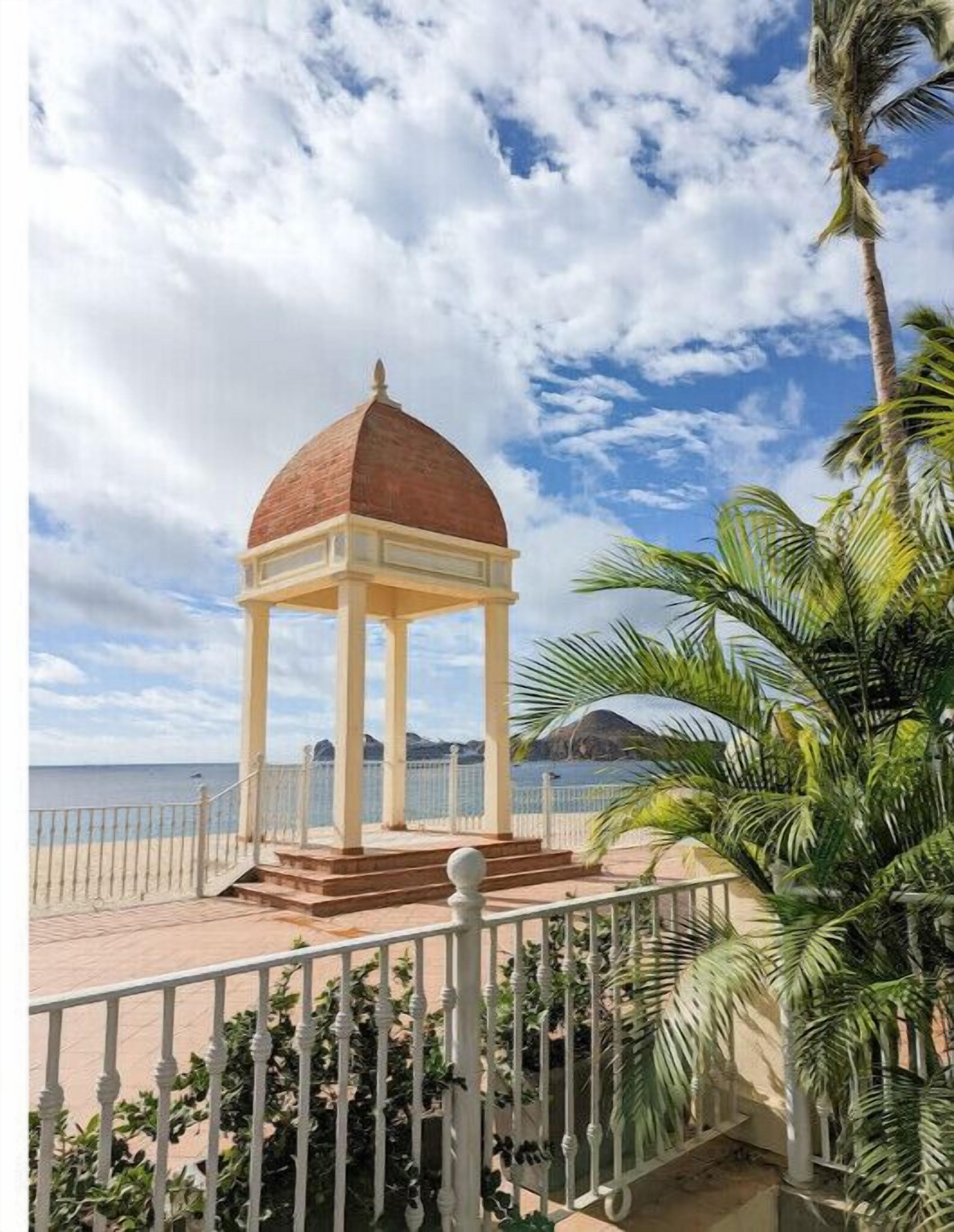 Ein Pavillon mit Kuppel, nahe einem Strand mit blauem Himmel und Palmen.