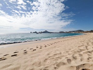 Ein sandiger Strand mit Fußspuren, die zum Wasser führen, ein klarer Himmel und ferne Berge.