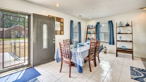 A dining area with a table set for four, surrounded by wooden chairs. There is a door with a glass panel, a window with blue curtains, and a shelf with decorative items. - Centrally Located Hampton Home: 6 Mi to Downtown! (Hampton)