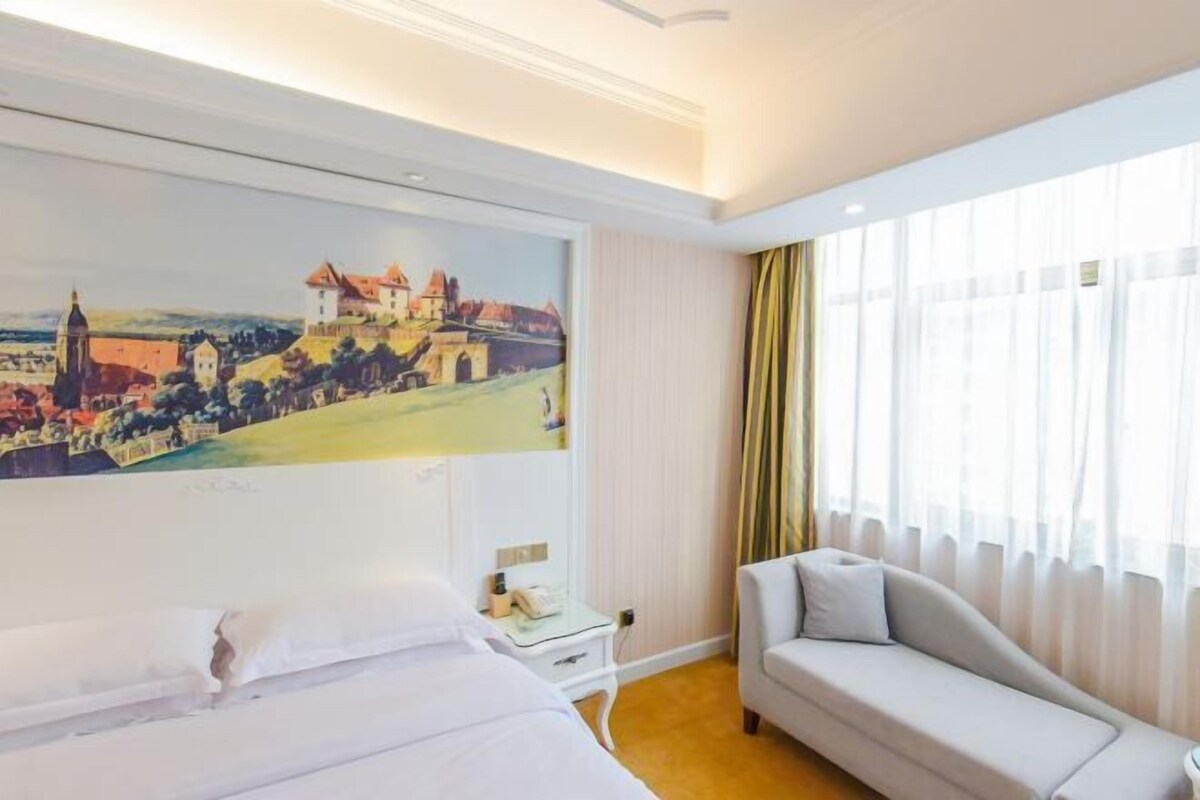 A hotel room with a bed, a sofa, a painting of a town on the wall, and a window with curtains.