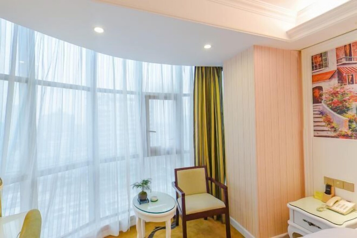 A hotel room with a large window, a small table with a plant, a chair, a bedside table with a phone, and a painting on the wall.