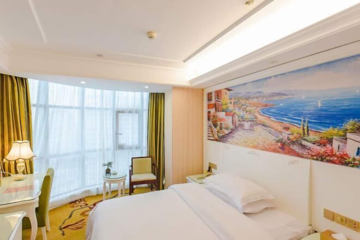 Une chambre d’hôtel avec un lit, un bureau, une chaise et un tableau représentant un paysage côtier.