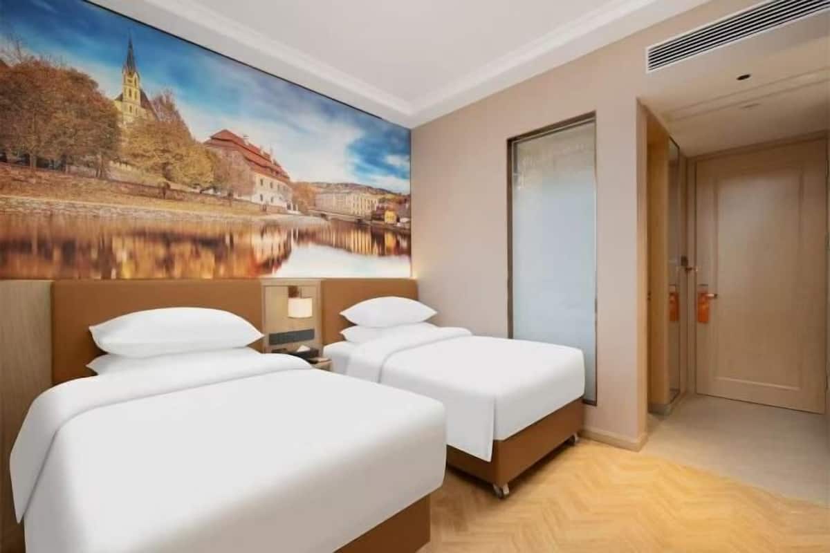 Une chambre d’hôtel avec deux lits, une grande fenêtre donnant sur un immeuble et des arbres, et une porte située à droite.