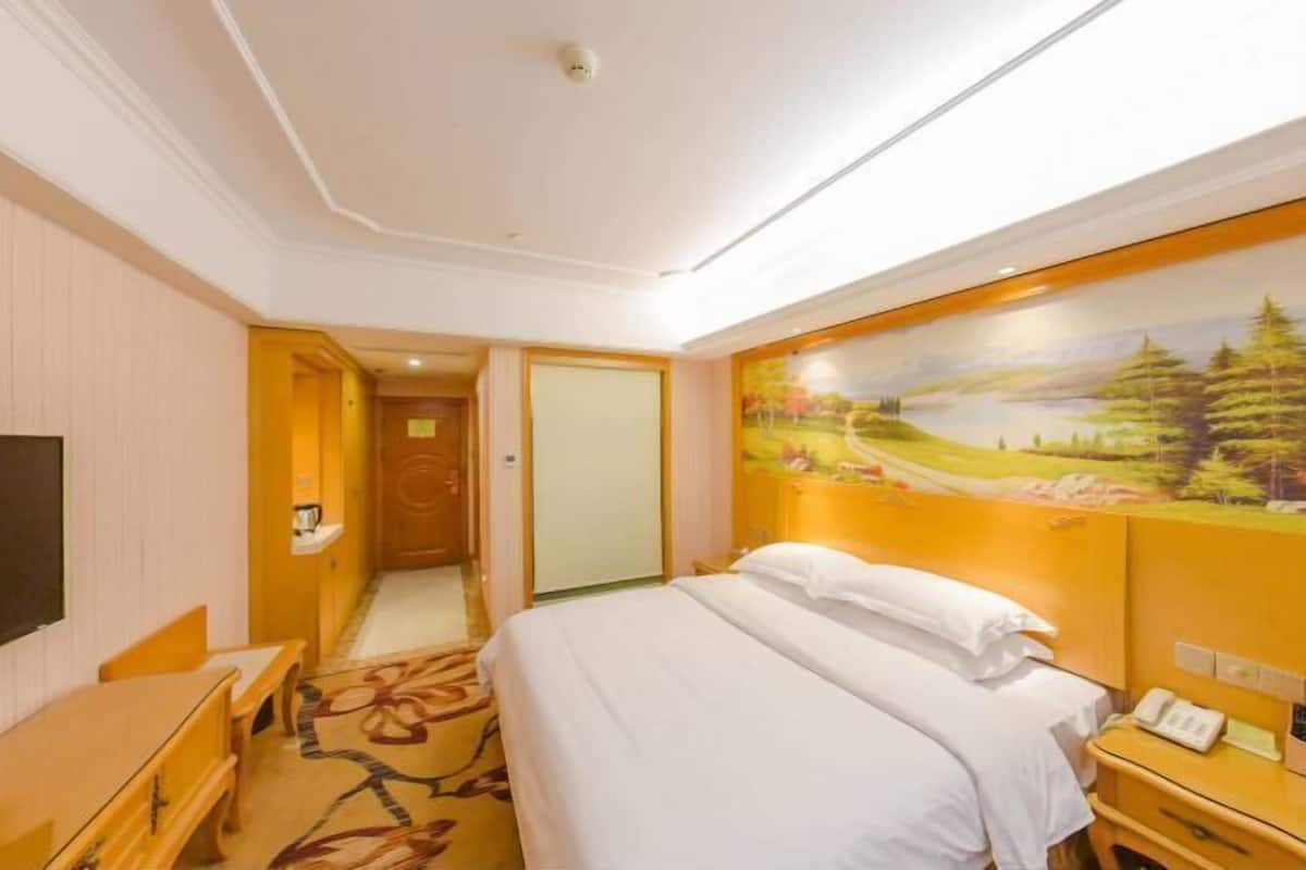 Une chambre d’hôtel avec un lit, un bureau, une chaise, un tableau au mur et une porte donnant sur une autre pièce.