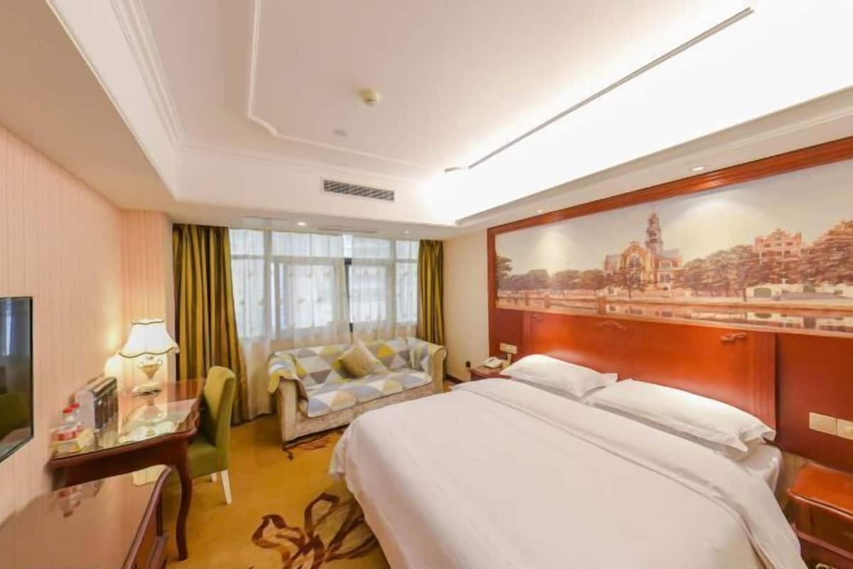 Une chambre d’hôtel avec un lit, un canapé, un bureau avec une lampe, un tableau au mur et une fenêtre avec des rideaux.
