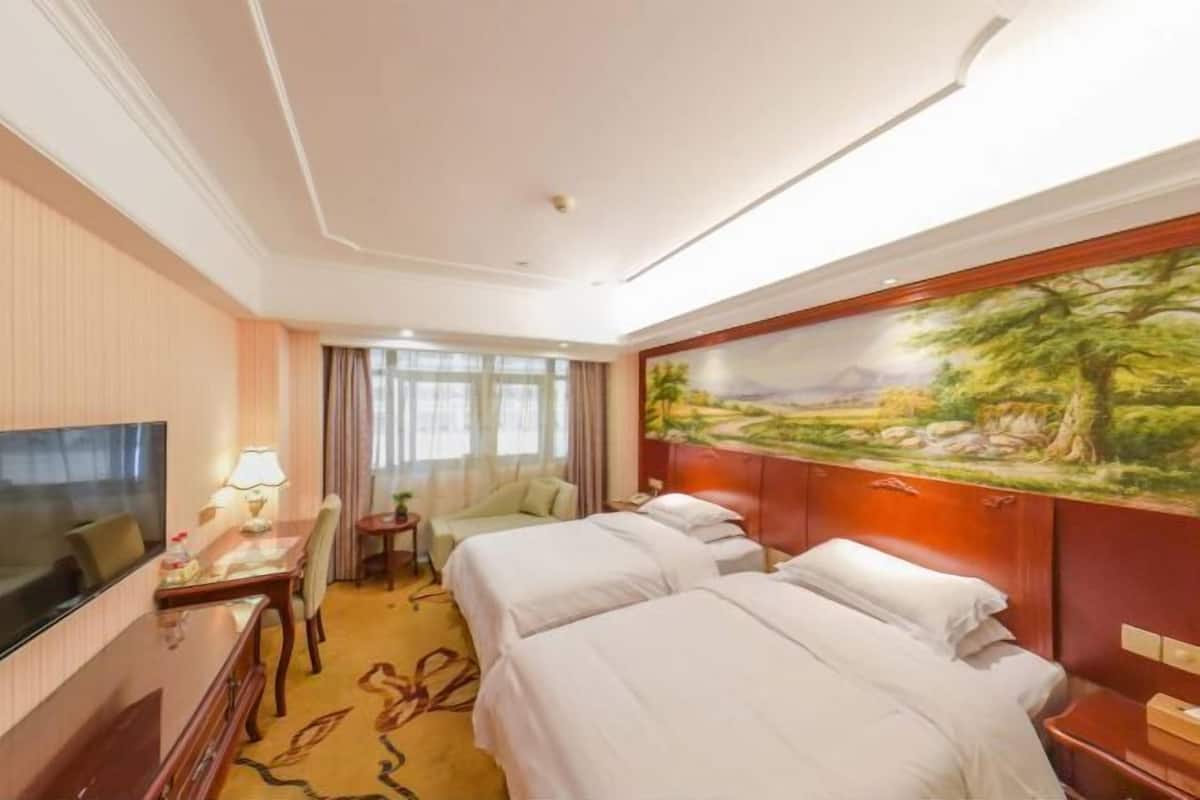 Une chambre d’hôtel avec deux lits, une télévision, un bureau avec une lampe, un tableau au mur et une vue sur l’extérieur.