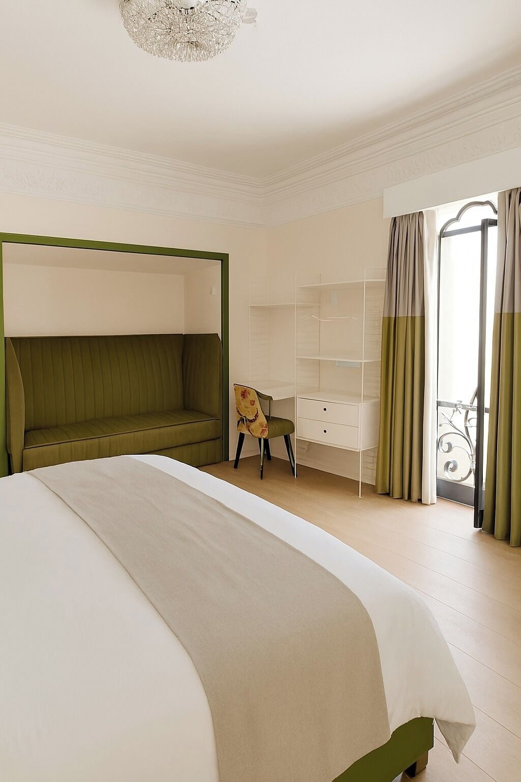 Un dormitorio con cama, un sofá verde, una silla, un aparador blanco y un balcón con cortinas.
