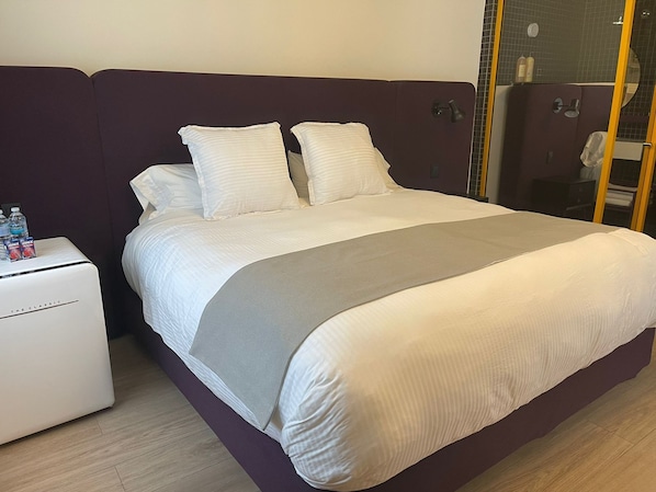 Un letto rifatto con lenzuola bianche e un dettaglio grigio, un comodino con bottiglie d'acqua e una testiera scura.