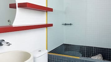 Un baño con inodoro blanco, lavamanos con encimera roja, cabina de ducha de cristal y suelo de baldosas.