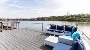 Eine Holzterrasse mit Sitzgelegenheiten im Freien und Blick auf einen See sowie Gebäude in der Ferne.