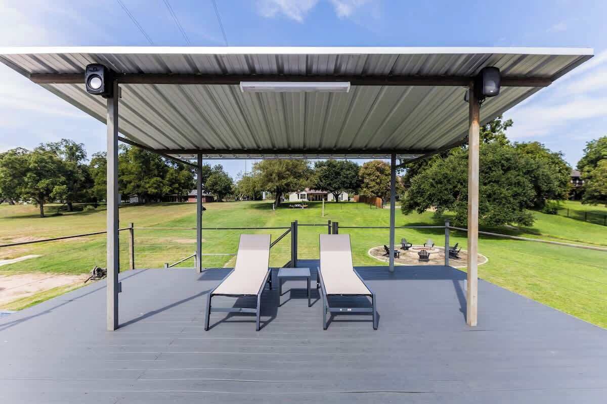 Un espacio de estar al aire libre con techo, que incluye dos sillones y una mesa, con vista a un parque con césped y árboles.