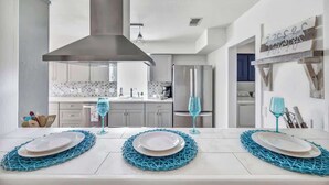 Eine moderne Küche mit weißen Schränken, einer Edelstahl-Dunstabzugshaube und einem Essbereich mit einem weißen Tisch, gedeckt für zwei Personen, mit blauen Platzsets.