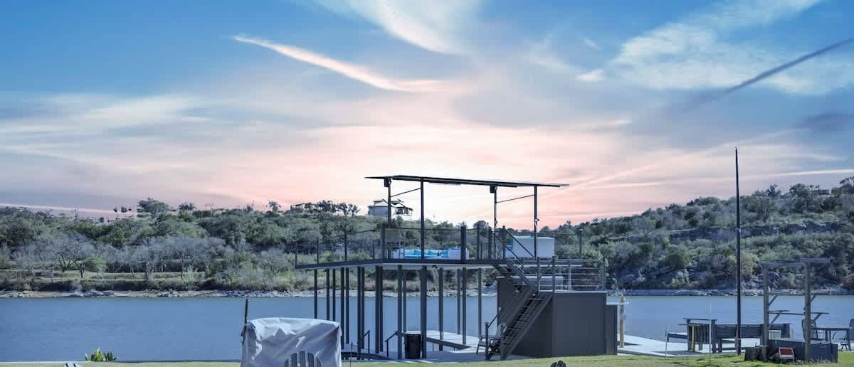 An outdoor seating area with Adirondack chairs and a fire pit, overlooking a lake with a boat slip structure in the background.