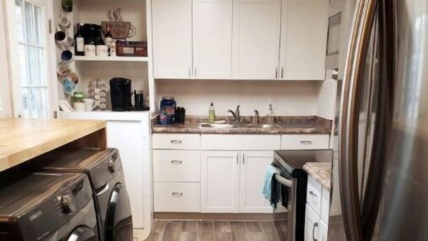 A compact kitchen with white cabinets, a stainless steel refrigerator, a washing machine, and a coffee bar sign.