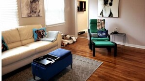 A living room with a beige sofa, a blue ottoman, and a green armchair. There are two large dog paintings on the walls.
