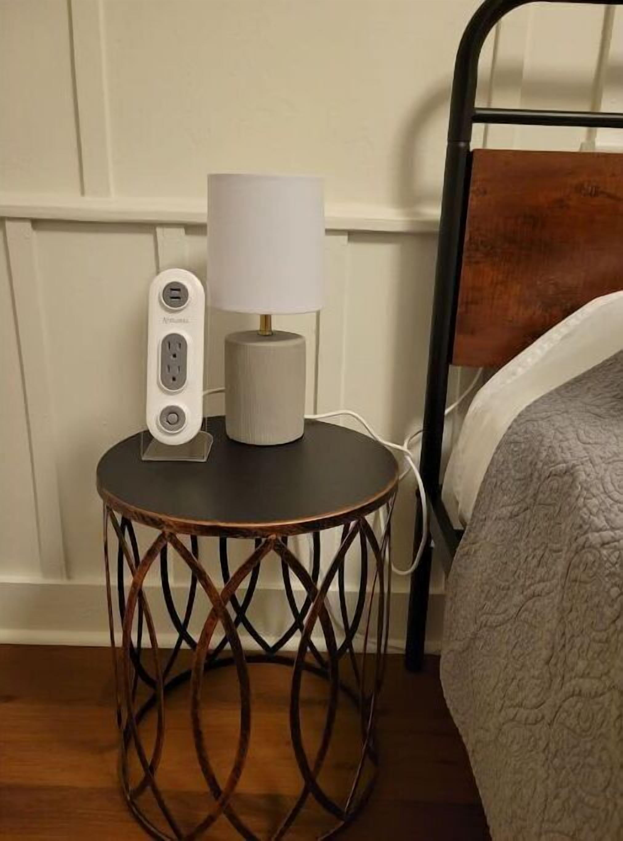 A small round table with a lamp and a power strip, positioned next to a bed in a room with wooden flooring and white walls.
