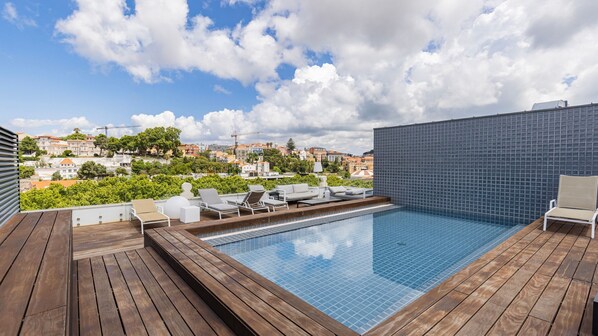A rooftop pool with wooden decking, lounge chairs, and a cityscape view.