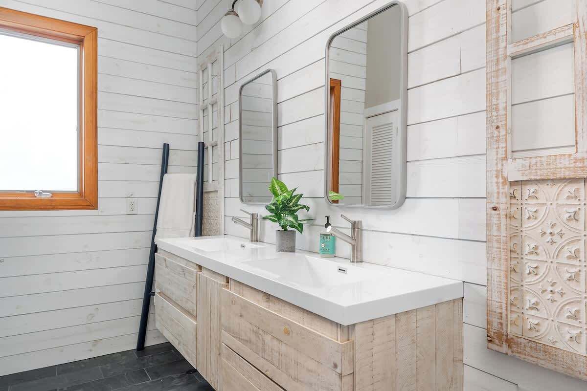 A bathroom with a white wooden wall, a double sink, two mirrors, a potted plant, and a decorative panel.