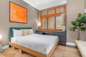 Une chambre à coucher avec un lit, un bureau, une fenêtre avec des stores, une plante et un tableau au mur.
