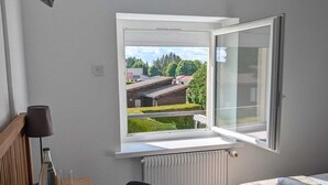 Hotelkamer met een bed, een bureau met lamp, een stoel en uitzicht op gebouwen door een open raam.