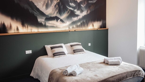 Een slaapkamer met een groot berglandschap op de muur, een bed met witte beddengoed, twee gestreepte kussens, en een opgerolde handdoek op het bed.