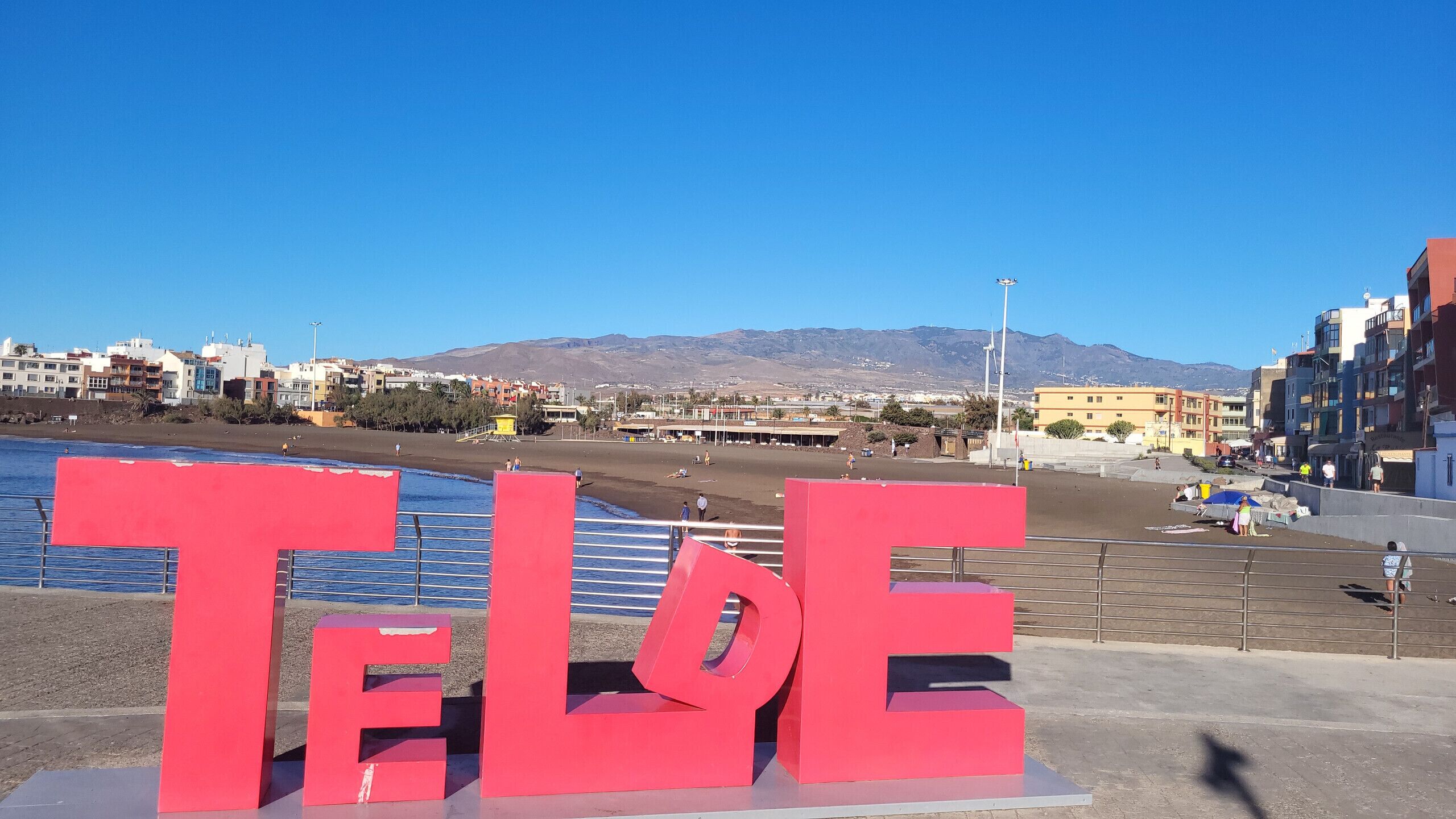 A beachside view with 'TELLE' sign, clear blue sky, and mountains in the background.