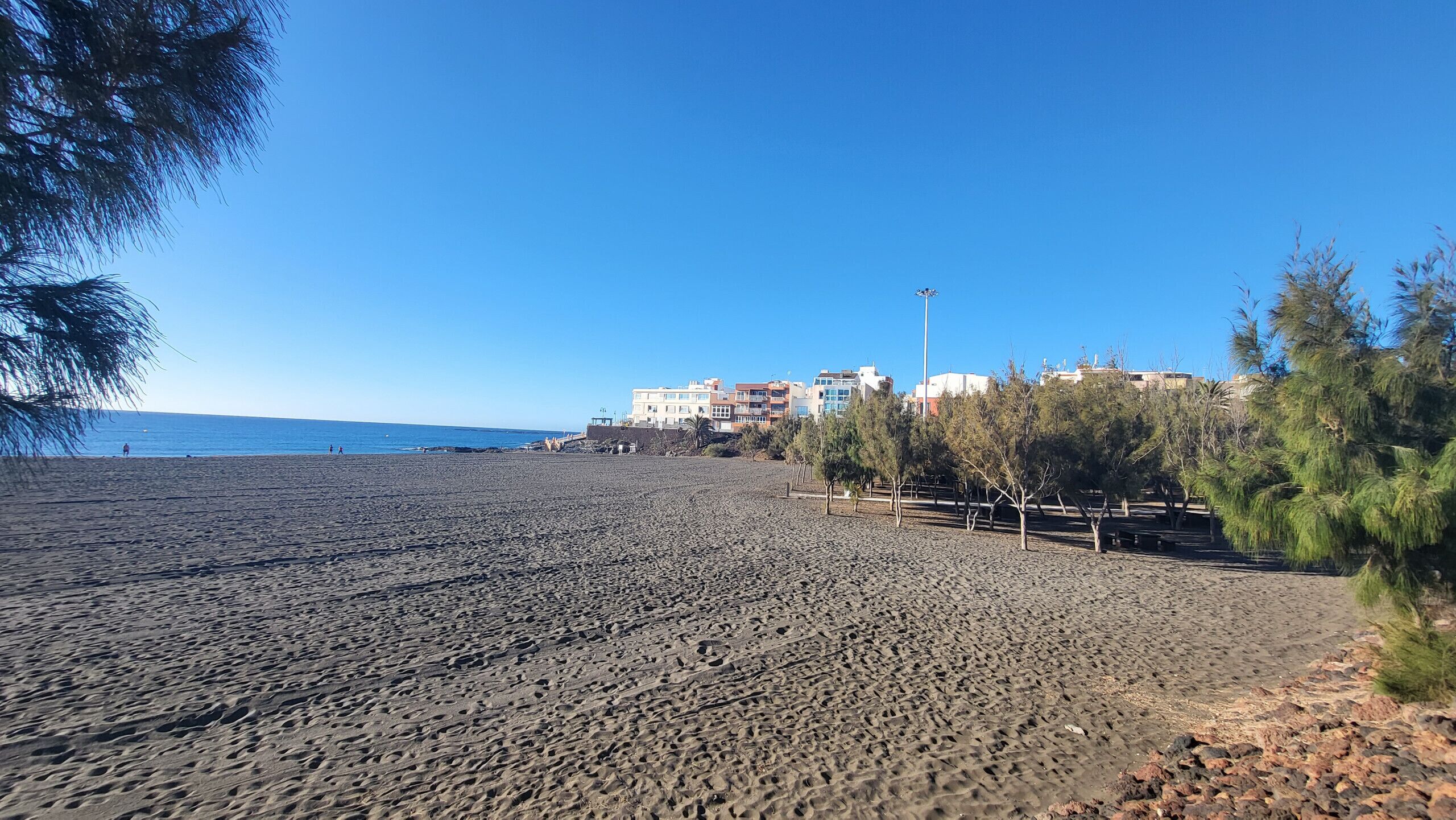 Een strand met rotsachtig terrein, een helderblauwe lucht en gebouwen op de achtergrond.