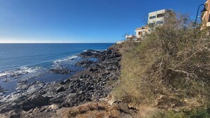 Zona costera con terreno rocoso, cielo azul despejado y un edificio moderno con balcones con vistas al mar.