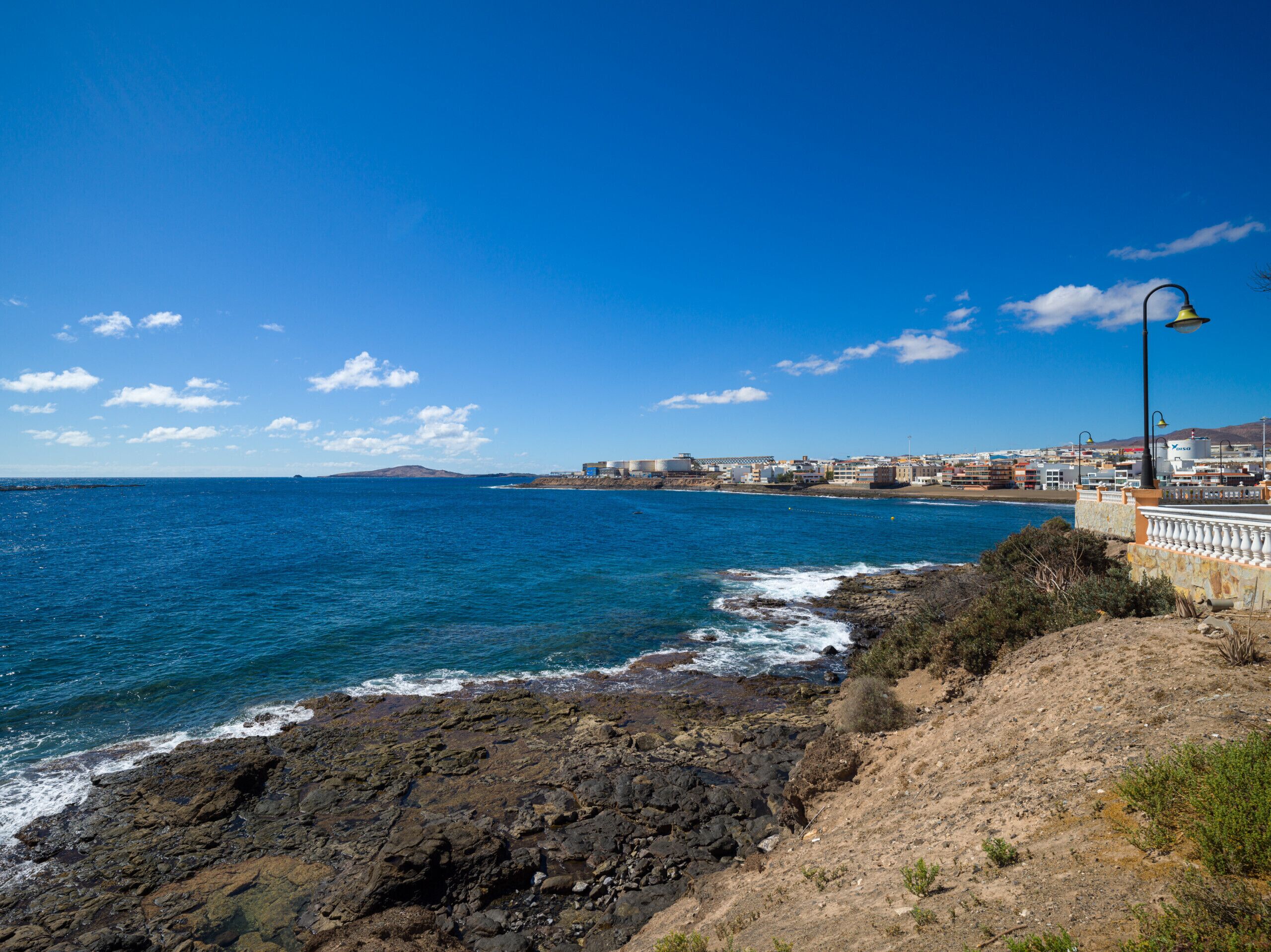 A coastal view with rocky terrain, a clear blue sky, and a distant town with buildings.