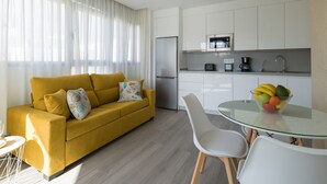 Een moderne woonkamer met een gele bank, een glazen eettafel met stoelen, en een keuken met witte kasten en een magnetron.