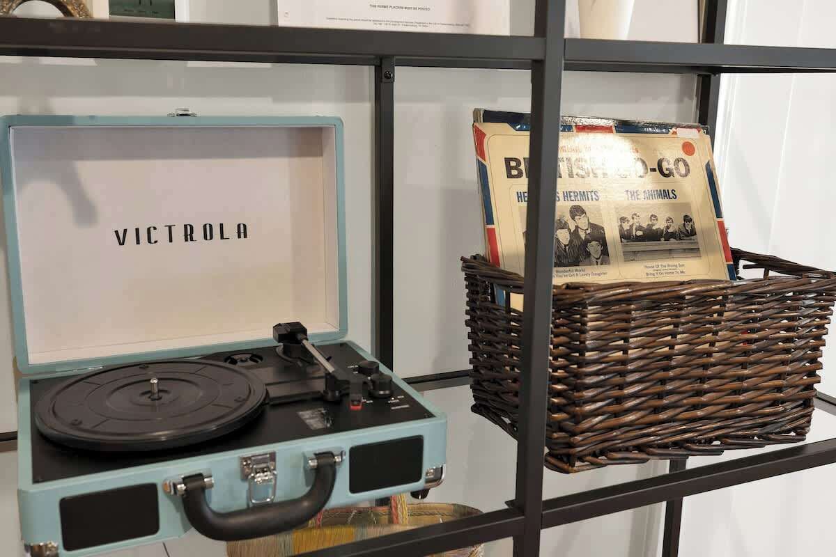 A vintage Victrola record player on a shelf, a wicker basket with magazines, and a framed newspaper.