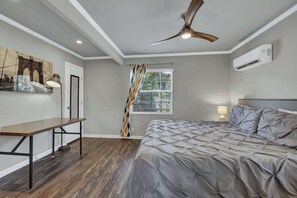 A bedroom with a bed, a desk, a ceiling fan, an air conditioner, a window with blinds, and a painting on the wall. - Luxury Mid Century Charmer Near Historic Main St (Fredericksburg)