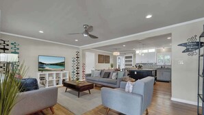 Ett rymligt vardagsrum med en grå soffa, ett soffbord och en tv. Längst bak finns ett kök med moderna vitvaror och en skylt med texten "Välkommen till Mulberry House".