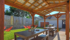Un patio al aire libre con mesas y sillas de madera, un área cubierta con techo de madera y un patio trasero cercado.