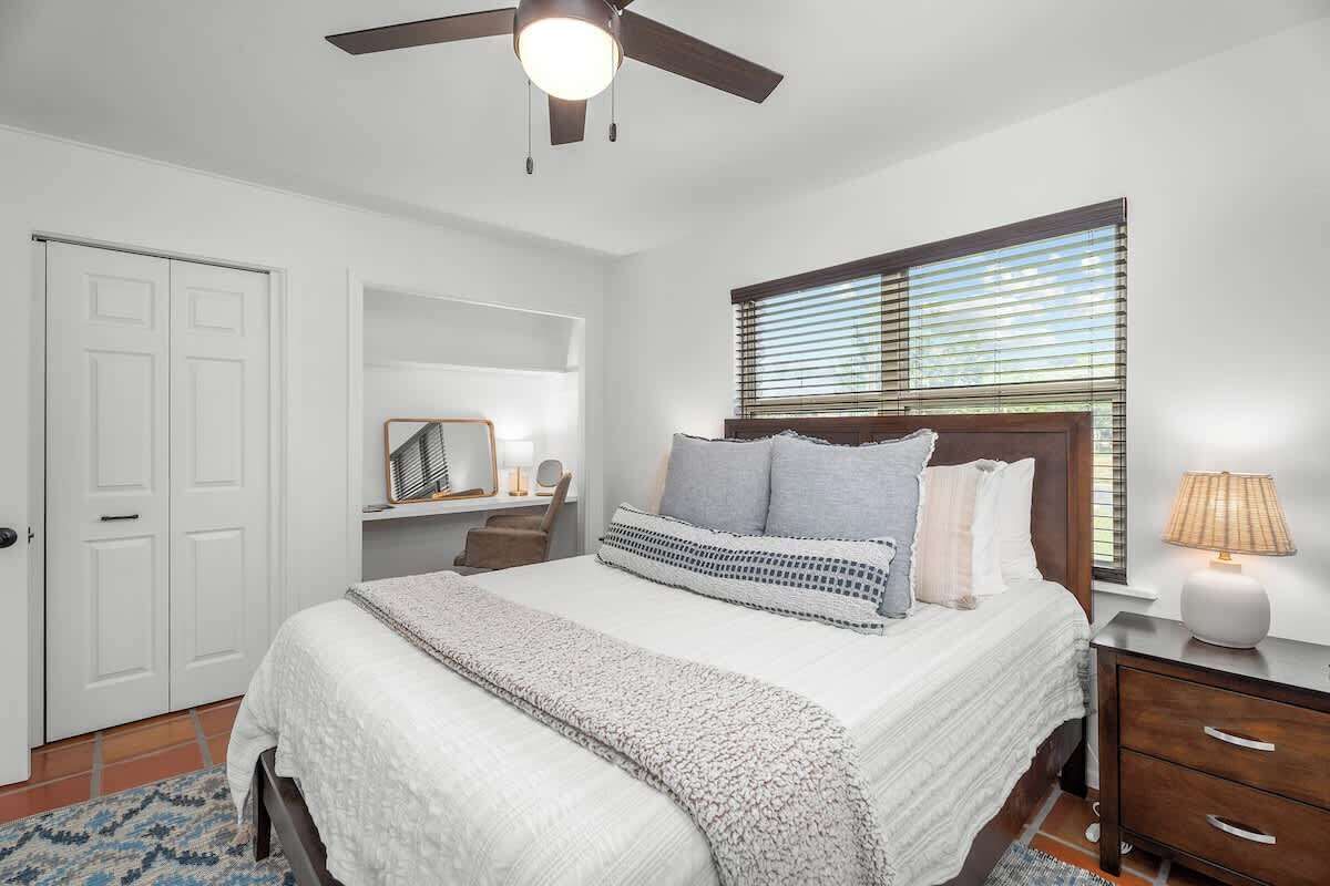 A bedroom with a bed, bedside table, lamp, window with blinds, ceiling fan, and a wardrobe.