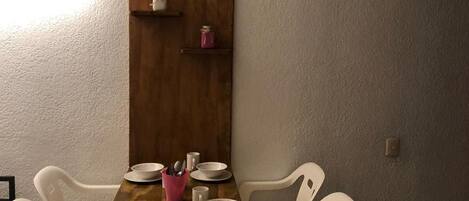 A dining area with a wooden table, white plastic chairs, and a white wall with a shelf.